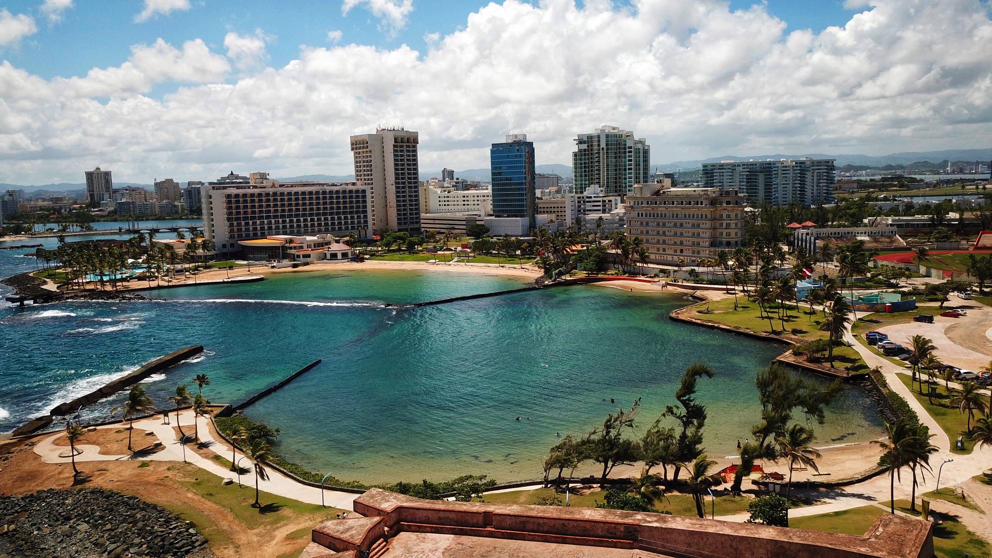 Aerial view of the Escambrón cove from above the Batería: turquoise water inside the breakwaters, the beach, and the Condado skyline behind